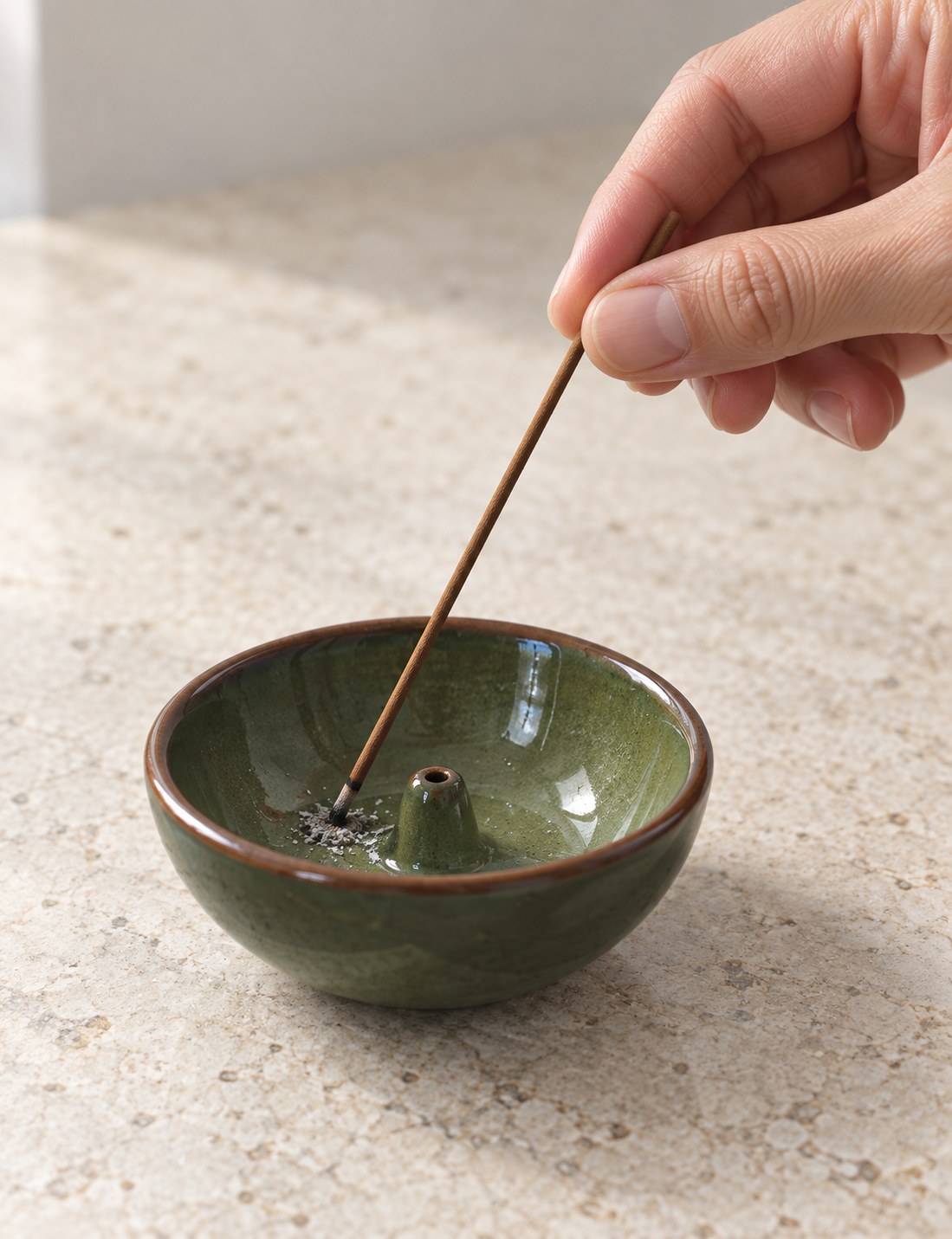 Hand gently pressing a charred incense tip against the inside of a green ceramic holder, with ash contained in the bowl
