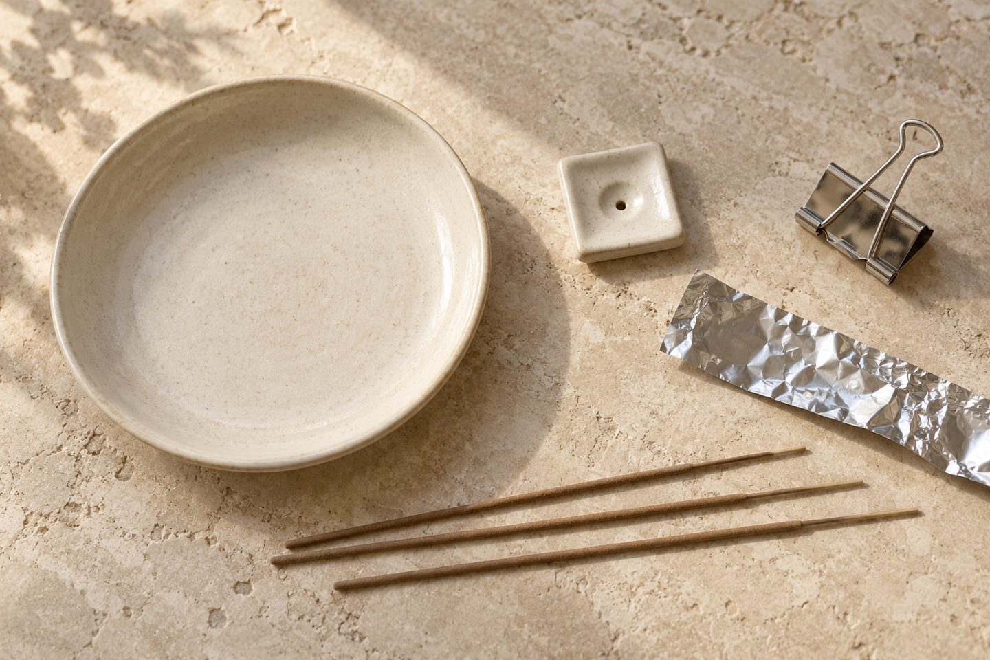A ceramic dish, small incense holder, metal binder clip, kitchen foil and loose unlit incense sticks on pale stone